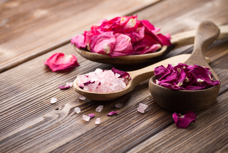 Bowls of rose petals and pink bath salts on wooden surface.